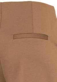 Gros plan d'un vêtement en tissu marron montrant une poche passepoilée horizontale avec des coutures propres et une texture lisse.