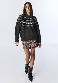 Maglione lavorato a maglia di un grigio scuro con motivi geometrici bianchi, abbinato a una minigonna scozzese rossa. Il modello indossa stivali alla caviglia neri e morbidi.