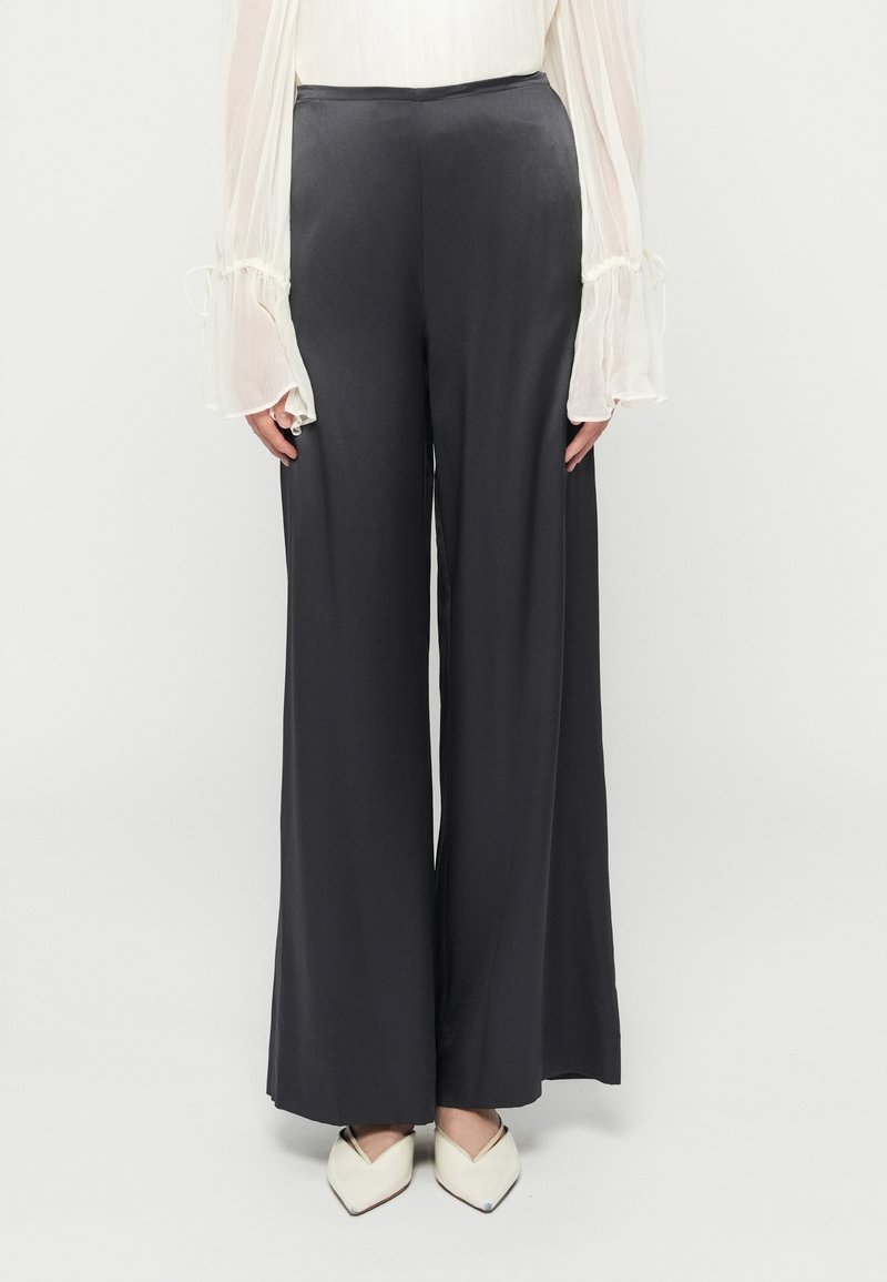 Femme portant un pantalon large noir, un chemisier blanc transparent à manches évasées et des chaussures blanches à bout pointu, debout devant un fond uni.