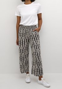 Personne portant des baskets blanches, un pantalon large noir et beige à motifs, et un t-shirt blanc à manches courtes uni, se tenant avec une main dans la poche.