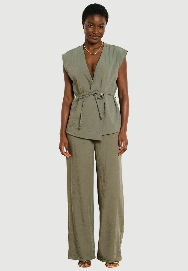 Donna che indossa un top verde oliva senza maniche con cintura da annodare e pantaloni larghi abbinati, in piedi davanti a uno sfondo semplice.