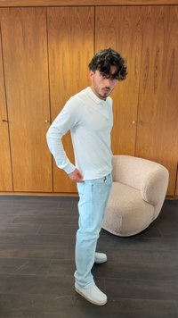 Polo shirt bianco a maniche lunghe, jeans blu chiaro e scarpe bianche. Posizione in piedi con una sedia beige testurizzata sullo sfondo.
