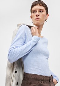 Svetlomodrý dlhý rukáv top vyrobený z jemného materiálu, s voľným strihom a okrúhlym výstrihom. V kombinácii s hnedými nohavicami s vysokým pásom a pruhovanou bundou.