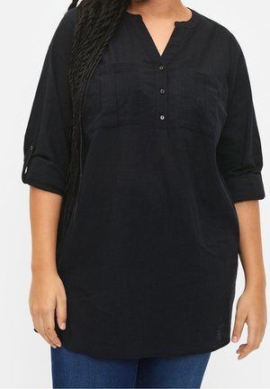 Kobieta ubrana w czarną bluzkę z długim rękawem, dekoltem w serek i kieszeniami na piersi oraz niebieskie jeansy, stojąca na jasnym, jednolitym tle.