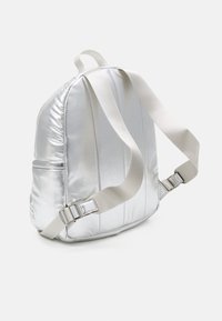 Mochila metálica plateada con un diseño acolchado, correas ajustables beige y un bolsillo lateral. Textura suave y forma elegante y redondeada.