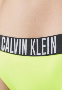 Neon gele bikini met een zwarte elastische tailleband waarop het 'CALVIN KLEIN' logo in het wit staat. Glad stof, halfhoge snit.