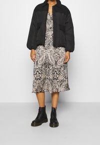 Personne portant une doudoune noire par-dessus une robe midi beige et noire à motif cachemire avec des bottes de combat noires à lacets.