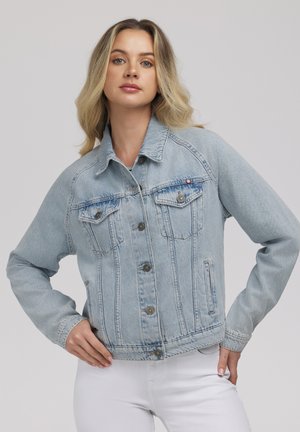 Młoda kobieta o blond włosach, ubrana w jasnoniebieską kurtkę dżinsową i białe spodnie, pozująca z rękami na biodrach na szarym tle.