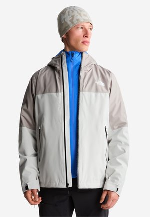 Mężczyzna w jasnoszarej i beżowej kurtce North Face, pod którą ma jaskrawo niebieską bluzę na zamek oraz beżową dzianą czapkę, patrzy w bok.