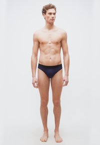 Νεαρός άνδρας όρθιος, ξυπόλητος, φοράει σκούρα μπλε εσώρουχα με μαύρη ζώνη στη μέση, μπροστά σε απλό λευκό φόντο.