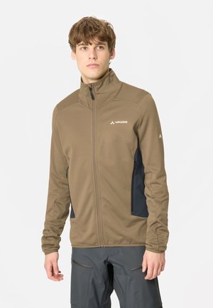 Jeune homme portant une veste marron zippée avec des panneaux noirs sur les côtés et un pantalon gris foncé, debout devant un fond blanc.