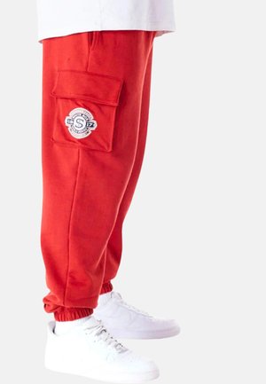 Red jogger pants with an elastic waistband and ankle cuffs, featuring a cargo pocket and a round logo patch on the side. Worn with white sneakers.