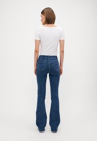 Jeans de mezclilla azules acampanados con una cintura de altura media y dos bolsillos traseros, combinados con una camisa blanca de manga corta y sin estampado. Textura suave.
