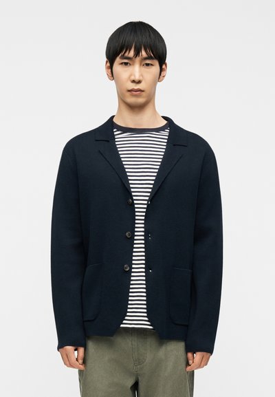 Jeune homme portant un cardigan bleu marine boutonné par-dessus une chemise rayée bleu marine et blanche, se tenant devant un arrière-plan blanc uni.
