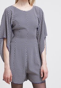 Tyrkysový a bílý vzorovaný overal s volánovými rukávy; má kulatý výstřih, viditelné knoflíky a pohodlný střih. Hladká textura látky.