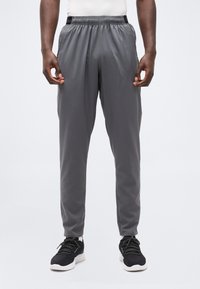 Άνδρας που φοράει γκρι αθλητικά παντελόνια και μαύρα αθλητικά παπούτσια, στέκεται με τα χέρια χαλαρά στα πλευρά, μπροστά σε ένα απλό λευκό φόντο.