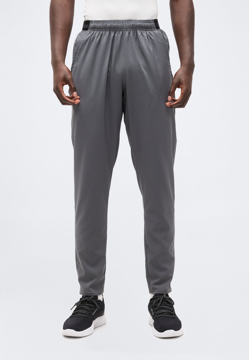 Άνδρας που φοράει γκρι αθλητικά παντελόνια και μαύρα αθλητικά παπούτσια, στέκεται με τα χέρια χαλαρά στα πλευρά, μπροστά σε ένα απλό λευκό φόντο.