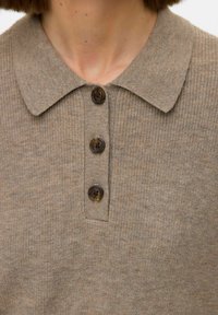 Bežs adīts polo krekls ar ribota tekstūra, ar norādītu apkakli un trim tumšām pogām centrā.