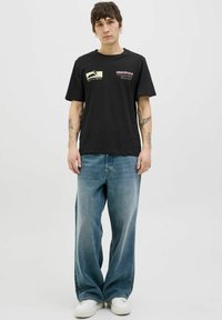 Zwarte katoenen t-shirt met grafisch logo, gecombineerd met lichtblauwe wijde jeans en witte sneakers, voor een effen achtergrond.