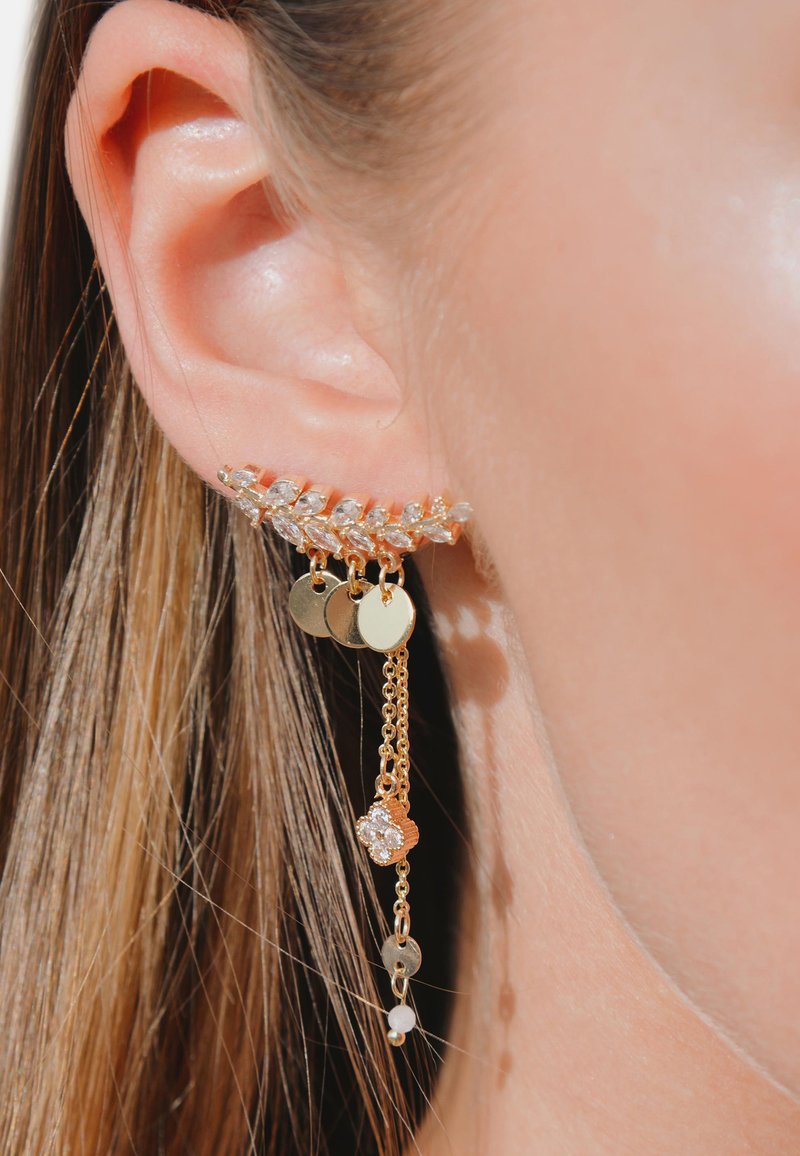 Boucle d'oreille en ton or avec un design floral orné de strass clairs, cercles dorés pendants et chaînes, sur un fond de peau naturelle.