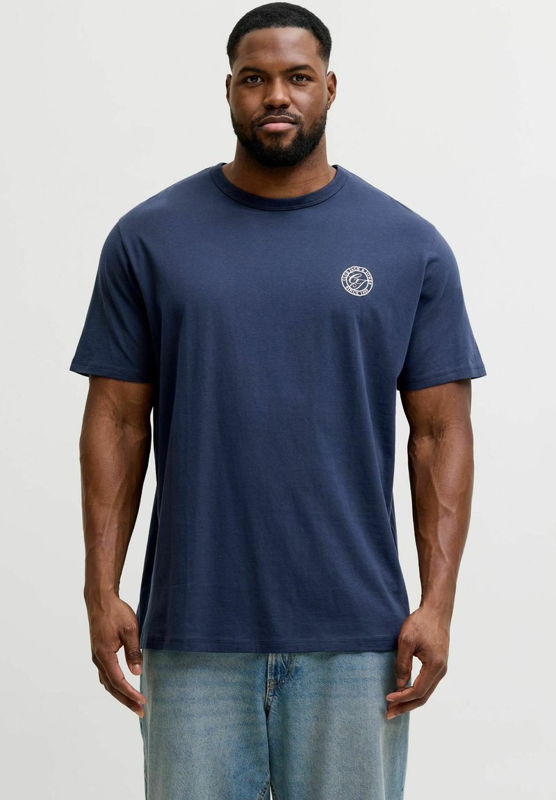 Mann mit kurzem Haar und Bart, trägt ein marineblaues T-Shirt mit einem kleinen weißen runden Logo auf der linken Brust und hellblaue Jeans.