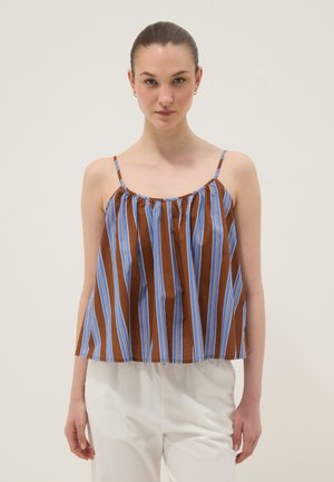 Donna che indossa un top ampio senza maniche a righe verticali marroni e blu con sottili spalline e pantaloni bianchi, in piedi davanti a uno sfondo semplice.