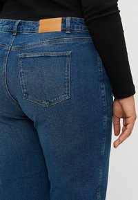 Jeans in denim blu scuro con vita media, dotati di una patta posteriore in pelle marrone e due tasche posteriori. Il tessuto ha una texture liscia.
