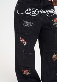 Pantalones de mezclilla negros que presentan múltiples bolsillos con parche y diseños bordados, incluyendo motivos de corazón y calavera, con el logotipo de "Ed Hardy".