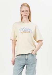 Bež pamučna majica kratkih rukava s plavo-smeđim natpisom "TIMBS Authentic", laganog kroja, u kombinaciji sa svijetloplavim trapericama.