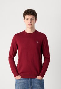 Bordeaux genser med rund hals laget av glatt stoff med lange ermer. Har ribbestrikkede mansjetter og nederkant, med en liten logo på brystet.