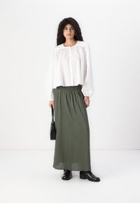 Chemisier blanc à boutons avec manches bouffantes, associé à une jupe maxi verte taille haute et des bottines noires à talon. Petite sac à main noir.