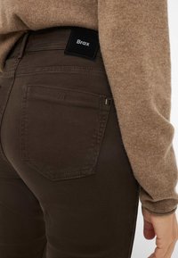Brune jeans lavet af denim med en tekstureret overflade, der har et sort Brax-label på livlinjen og en baglomme med syet kant.