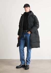 Black padded coat with a hood, black turtleneck, blue denim jeans, and black leather boots. Model wears a black cap. Neutral background.