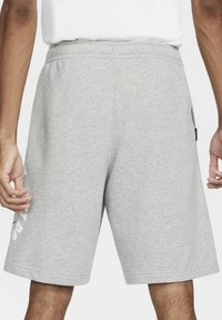 Pantalones cortos atléticos grises de algodón con cinturilla elástica, bolsillos laterales y detalle impreso en blanco en la pierna izquierda. Textura suave y lisa.
