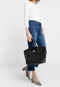Borsa tote nera con stampa a coccodrillo, dalla forma strutturata, con manici lisci e dettagli in metallo. Abbinata a jeans blu e ballerine con motivi.