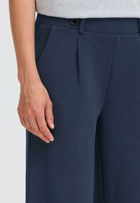 Navy trousers featuring a smooth texture, front pleats, and a side pocket. A black button is visible at the waist. Hand resting on the fabric.