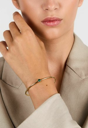 Bracciale rigido in metallo dorato con un'accento centrale in pietra verde. Indossato su una mano con un outfit tono neutro e una texture della pelle liscia.