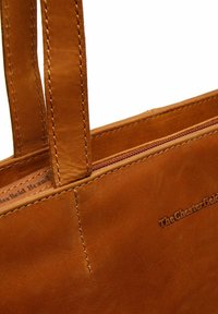 Bruine leren tote bag met een gladde textuur, voorzien van een rits aan de bovenkant en twee stevige handvaten. Logo in de zijkant geperst. Minimale stikseldetails.