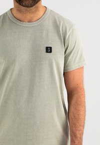 Lichtgroene T-shirt van zacht katoen, met een ronde halslijn, korte mouwen en een klein zwart vierkant logo op de borst.