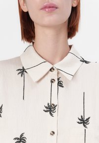 Lys beige skjorte med svart palmebroderi, knapp-opplegg foran og klassisk krage. Teksturert stoff med en subtil glans.