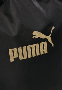 Sac en tissu noir avec un logo Puma en paillettes dorées et du texte. La surface est lisse avec un léger éclat, mettant en avant des éléments de design minimalistes.