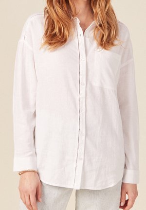 Kvinde iført en løs hvid skjorte med knapper og brystlomme, kombineret med lysegrå bukser, hendes hår er løst og hun har et armbånd på håndleddet.