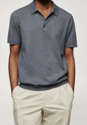 Person trägt ein graues Poloshirt mit kurzen Ärmeln und zwei Knöpfen sowie beige Hose, die Hände in den Taschen ruhend vor neutralem Hintergrund.