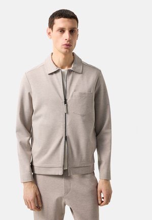 Mężczyzna w jasnobeżowej kurtce na zamek z kieszenią na piersi i pasujących spodniach na jednolitym jasnym tle.