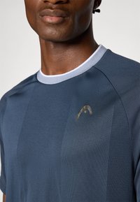 Camiseta atlética azul oscura con un acabado texturizado, que presenta un collar blanco y un sutil logo dorado en el pecho. Mangas cortas, tejido ligero.