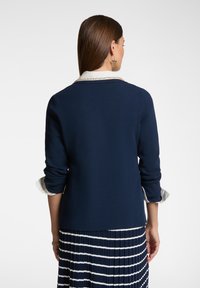 Maglione blu navy con una camicia bianca col collo sotto, dotato di maniche arrotolate e un finish a maglia testurizzato. La vista da dietro mostra linee pulite.