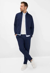 Chemise bleu marine à boutons portée sur un T-shirt blanc, associée à un pantalon bleu marine et des baskets blanches. La chemise présente deux poches poitrine et une coupe décontractée.