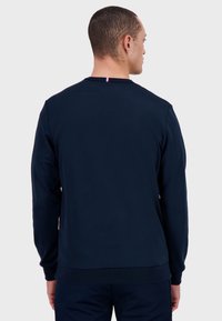 Felpa navy con collo a giro e polsini a costine. Presenta un piccolo dettaglio tricolore nella parte posteriore del collo. Tessuto morbido e textured.