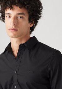 Camisa negra de botón con tejido suave, que presenta un cuello, un sutil detalle de logo y puños estándar, posada sobre un modelo.