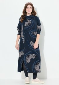 Mörkblå klänning med långa ärmar, prydd med ett fläktmönster i beige och ljusblått. Den har en sidoklyvning och är kombinerad med ljusa sneakers.